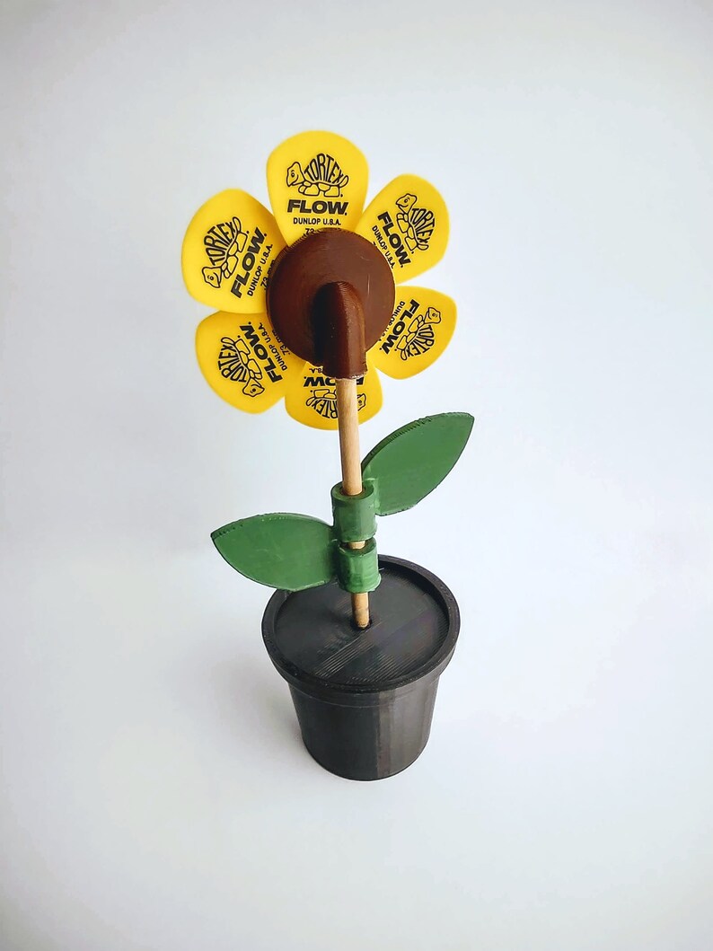Puede incluir: Una flor amarilla hecha de p&uacute;as de guitarra con el texto "FLOW" y "DUNLOP U.S.A." en cada p&uacute;a. La flor est&aacute; en un palo marr&oacute;n y en una maceta negra con hojas verdes.