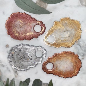 Pode incluir: Quatro pratos de anel de resina em diferentes cores e padrões. Os pratos têm a forma de ovais irregulares com um furo no centro. As cores são dourado, prateado, cobre e rosa.