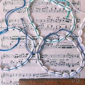 May include: Three white shell necklaces with blue, green, and white string. The necklaces are laid out on a sheet of music with the text "C. M. von WEBER, Op. 65" printed at the top.