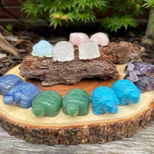 Puede incluir: Una colección de pequeñas figuritas de tortugas talladas en diferentes piedras preciosas, incluyendo amatista, cuarzo rosa, cuarzo transparente, turquesa y aventurina verde. Las figuritas están dispuestas en círculo sobre una rodaja de madera.