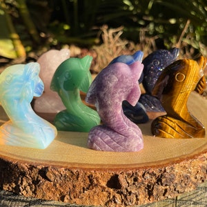 May include: A selection of carved gemstone dolphin figurines in various colours, including opalescent, pink, green, purple, blue, and tiger's eye. The dolphins are displayed on a round wooden slice, showcasing their detailed craftsmanship.