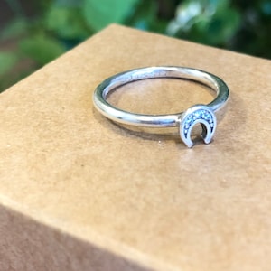 Puede incluir: Un anillo de plata con un diseño de herradura con pequeñas piedras transparentes. El anillo está sobre una superficie marrón.