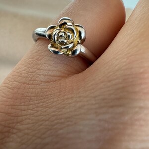 Puede incluir: Un anillo de plata con un diseño detallado de rosa. Los pétalos de la rosa tienen un centro dorado, creando un efecto de dos tonos. El anillo se muestra en un dedo, destacando la joyería floral.