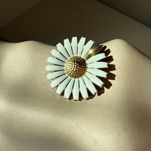 May include: A gold daisy brooch with white enamel petals and a textured gold centre. The brooch is a floral design, with a classic and elegant aesthetic. The brooch is a jewellery item.