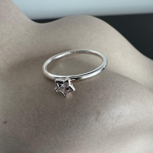 Puede incluir: Un anillo de plata con un dije en forma de estrella. El anillo presenta una banda delicada y una estrella de cinco puntas con una piedra clara y brillante en el centro. El anillo se muestra sobre una superficie de tono neutro.