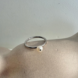 Puede incluir: Un anillo de plata con una sola perla blanca y redonda. El anillo tiene una banda texturizada y la perla está engastada en la parte delantera. El anillo se muestra sobre una superficie de tono neutro.