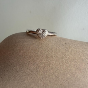 Puede incluir: Un anillo de plata con una piedra central en forma de corazón texturizado. La banda del anillo es delgada y pulida, y el corazón es de plata clara y texturizada. El anillo está sobre un fondo neutro.