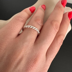 Puede incluir: Un anillo de plata con un delicado diseño de cadena. El anillo se lleva en un dedo con esmalte de uñas rojo.