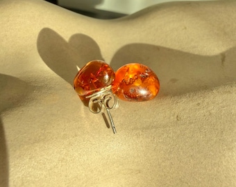 May include: A pair of amber stud earrings. Each earring features a rounded, polished amber stone with visible internal structures. The amber stones are set on silver posts with butterfly backs. The earrings are displayed on a neutral-toned surface.