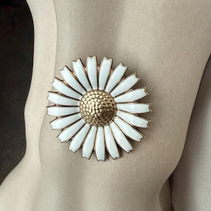 May include: A large daisy brooch with white enamel petals and a textured gold-tone centre. The brooch has a gold-tone border and is a classic floral accessory. The brooch is shown on a neutral background.