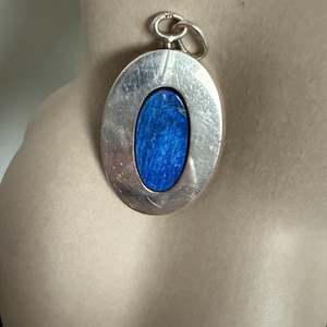 Puede incluir: Colgante ovalado de plata con una incrustación de piedra azul vibrante. El colgante tiene un acabado de metal cepillado texturizado y un pequeño lazo en la parte superior para una cadena. La piedra azul contrasta con la plata.