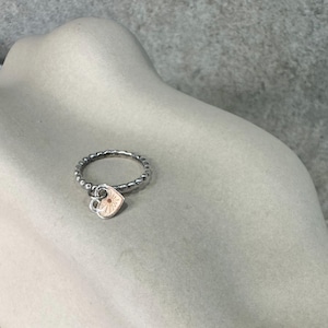 Puede incluir: Un anillo de plata con un pequeño dije en forma de corazón. El dije en forma de corazón tiene un delicado diseño floral en el centro.