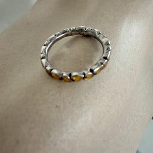 May include: A silver-toned ring featuring a continuous band of small, heart-shaped cutouts. Each heart-shaped section is filled with a warm, amber-coloured inlay. The interior of the band displays visible stamped markings along the metal surface.