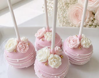 Könnte beinhalten: Vier runde Cake Pops an weißen Stielen, jeweils mit hellrosa Glasur überzogen. Die Cake Pops sind mit kleinen, aufgespritzten weißen und rosa Rosen aus Zuckerguss, feinen weißen Glasurfäden und winzigen goldenen Zuckerperlen verziert.