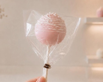 May include: A spherical cake pop coated in light pink icing with white drizzle and small white sprinkles, wrapped in clear plastic packaging on a white stick. The treat features a smooth finish and is held upright by a thin white paper stick.