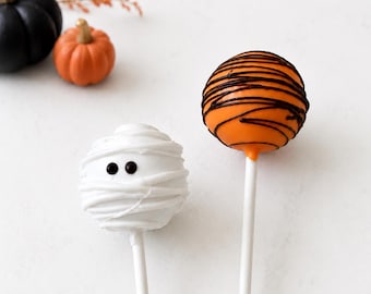 Könnte beinhalten: Zwei Cake-Pops an weißen Stielen. Einer ist weiß mit einem mumienartigen Wickelmuster und zwei kleinen schwarzen Augen. Der andere ist orange mit dünnen, dunkelbraunen Schokoladenstreifen. Ein schwarzer und ein orangefarbener Kürbis liegen daneben.