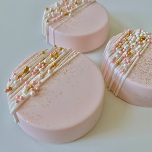 May include: Three pink round cookies decorated with white and gold drizzled icing and pink and gold sprinkles.