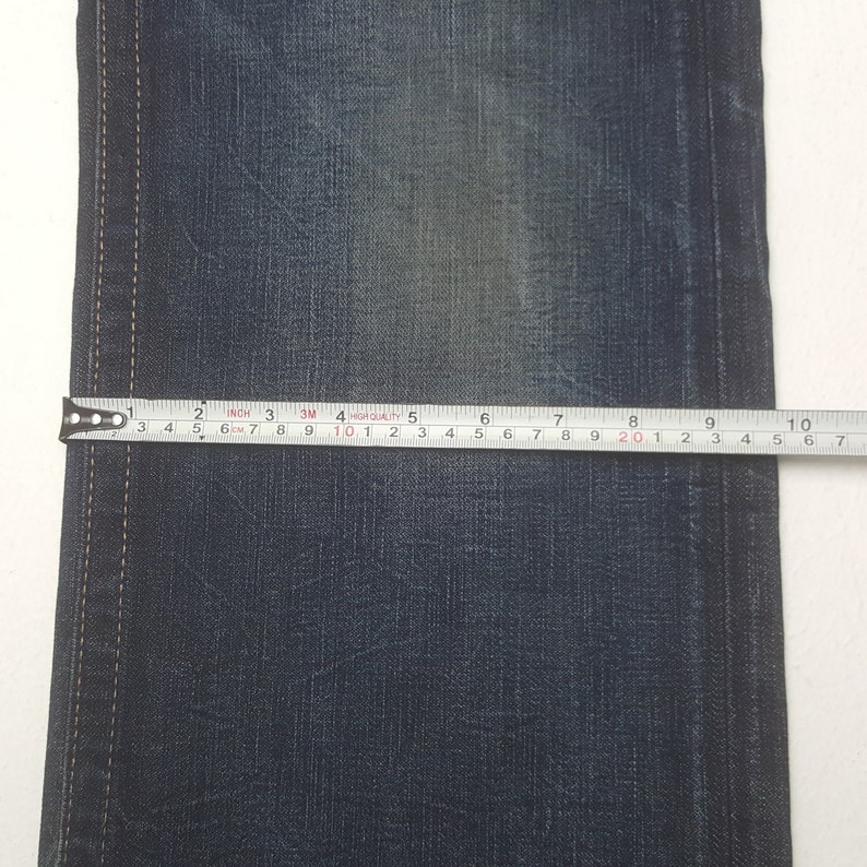 May include: Close-up of blue denim fabric with a measuring tape showing a width of approximately 25 cm. The tape measure is positioned horizontally across the fabric, indicating a measurement of the jeans.
