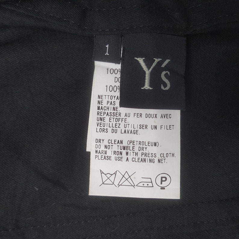 May include: Close-up of a black garment tag with white text and the brand logo "Y's". The tag includes care instructions in English and French, such as "Dry Clean" and "Do Not Tumble Dry".
