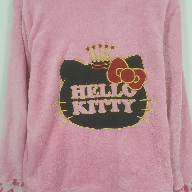 May include: Pink long-sleeved pajama top with a Hello Kitty graphic. The graphic features Hello Kitty's face with a crown and bow, and the words "HELLO KITTY". The cuffs have a pink and white animal print.