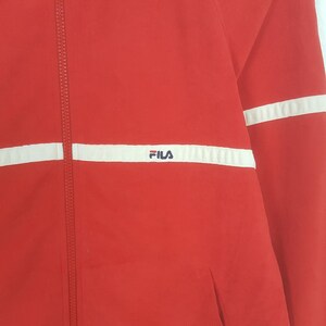 May include: Red athletic jacket with white stripes and the FILA logo. The jacket has a full zipper and a pocket. The jacket is made of a lightweight material.