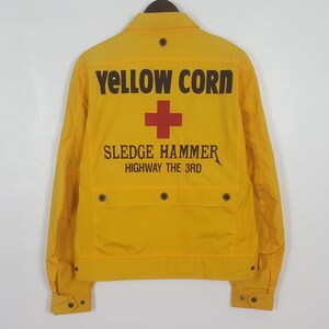 Może przedstawiać: Jasnożółta kurtka z czarnym napisem "YELLOW CORN" i "SLEDGE HAMMER" na plecach. Nad napisem znajduje się czerwony krzyż. Kurtka posiada dwie przednie kieszenie i zapięcia na guziki.