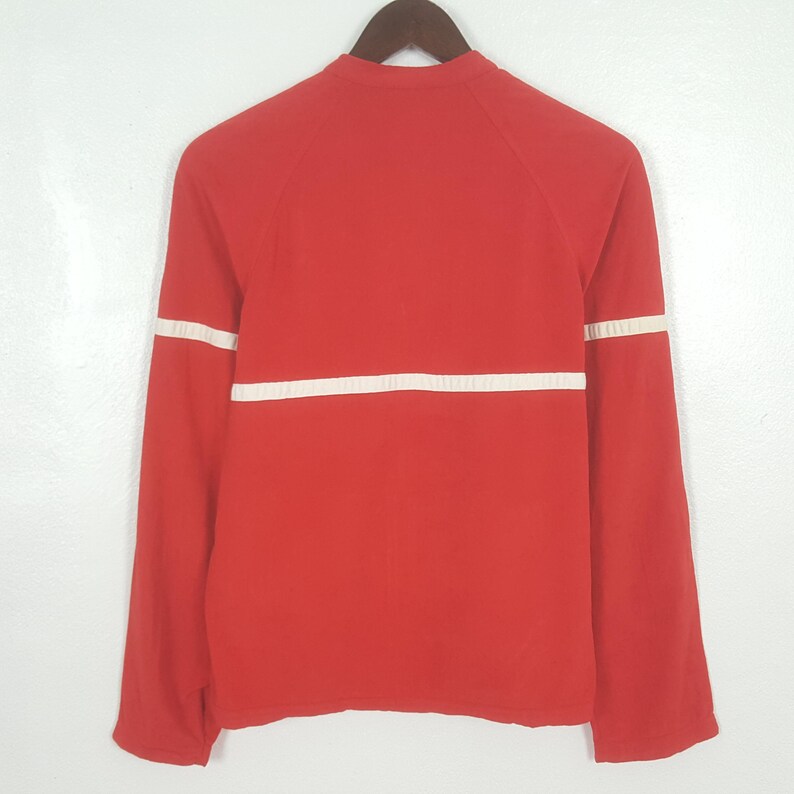 May include: A red long-sleeved shirt with a white horizontal stripe across the chest and white stripes on the sleeves. The shirt is hanging on a wooden hanger against a white background. The shirt has a crew neck.