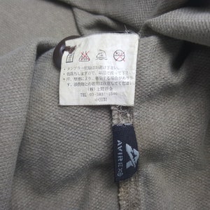 Puede incluir: Primer plano de una tela beige con una etiqueta y un bot&oacute;n marr&oacute;n. La etiqueta tiene texto japon&eacute;s y s&iacute;mbolos de cuidado. Tambi&eacute;n es visible una etiqueta negra con la marca "AVIREX".