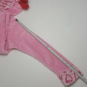 May include: A pink plush hooded robe with a flamingo design. The robe features a hood with a leopard print pattern and two red pom-poms. The sleeve is measured at 23 inches long.