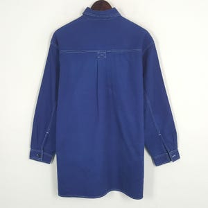 May include: A long-sleeved, cobalt blue shirt with white stitching details. The shirt features a classic collar, buttoned cuffs, and a back yoke with an X-shaped reinforcement stitch. The garment is displayed on a wooden hanger.