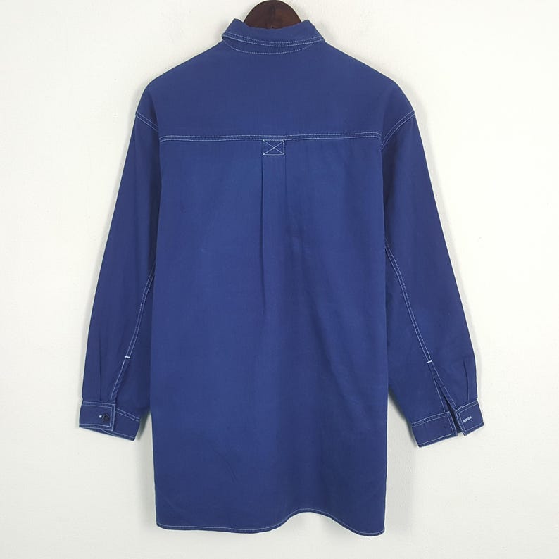 May include: A long-sleeved, cobalt blue shirt with white stitching details. The shirt features a classic collar, buttoned cuffs, and a back yoke with an X-shaped reinforcement stitch. The garment is displayed on a wooden hanger.