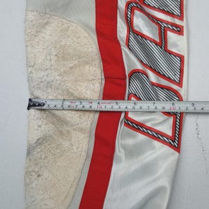 Peut inclure: Gros plan sur une section d'un pantalon de sport. Le pantalon est principalement gris clair avec une bande rouge et du texte noir et blanc. Un m&egrave;tre ruban indique une mesure d'environ 23 cm.