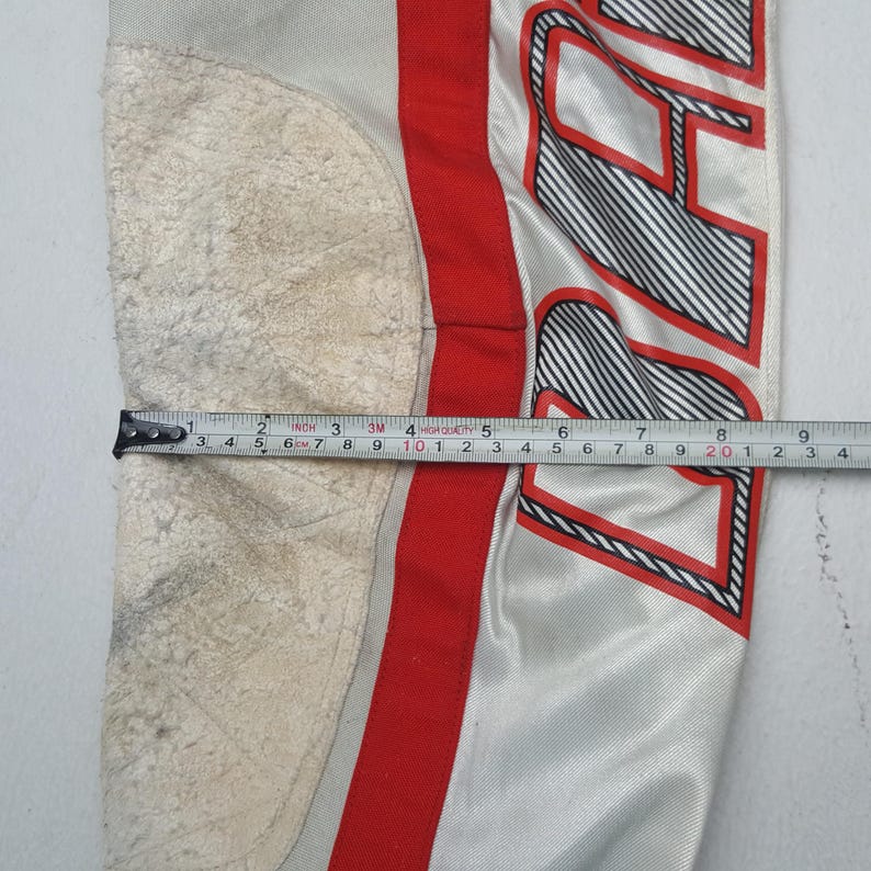 Peut inclure: Gros plan sur une section d'un pantalon de sport. Le pantalon est principalement gris clair avec une bande rouge et du texte noir et blanc. Un m&egrave;tre ruban indique une mesure d'environ 23 cm.