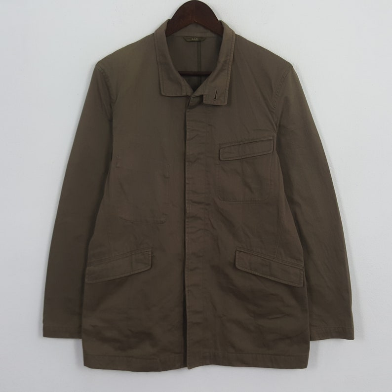 Puede incluir: Chaqueta verde oliva con cuello, dos bolsillos delanteros y cierre de botones. La chaqueta est&aacute; colgada de una percha de madera sobre un fondo blanco. La chaqueta tiene mangas largas.