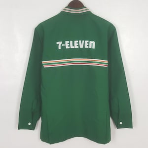 Op de afbeelding: Groene jas met lange mouwen en de tekst "7-ELEVEN" in witte letters op de rug. De jas heeft een horizontaal streepontwerp in rood, oranje en wit. De kraag heeft een gestreept ontwerp in groen, wit en oranje.