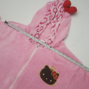 May include: Pink plush hooded onesie with a white zipper and a Hello Kitty graphic. The hood features a pink leopard print pattern and red bow accents. The onesie measures approximately 20 inches in length.
