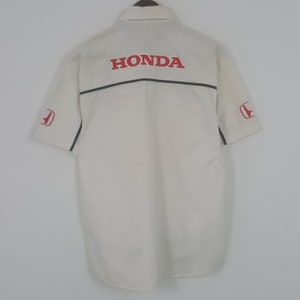 Peut inclure: Chemise crème à manches courtes avec le mot "HONDA" en rouge dans le dos. La chemise a une bande noire et un logo Honda rouge sur la manche gauche. La chemise est boutonnée.