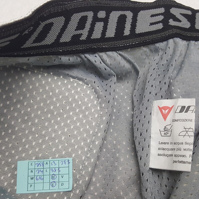 Peut inclure: Gros plan sur un v&ecirc;tement en maille grise avec le nom de la marque "DAINESE" en noir et gris en haut. Une &eacute;tiquette blanche avec des instructions de lavage et un autocollant bleu et vert avec des chiffres et des lettres sont visibles.
