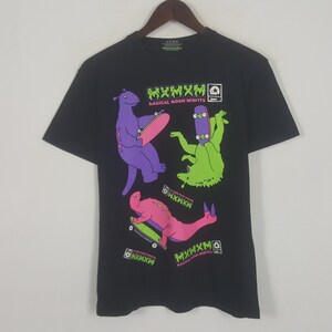 May include: Black t-shirt featuring three cartoon dinosaurs skateboarding. The dinosaurs are purple, green, and pink, with matching skateboards. The shirt has the text "MXMXM MAGICAL MOSH MISFITS" in green and white.