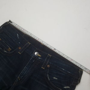 May include: Dark blue denim jeans with yellow stitching and a silver button. A measuring tape shows the jeans are approximately 48 cm across the waist. The jeans have a classic five-pocket design and a distressed look.
