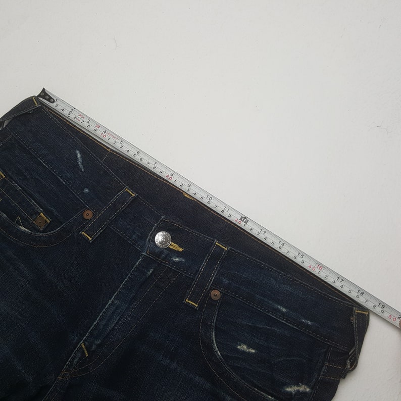 May include: Dark blue denim jeans with yellow stitching and a silver button. A measuring tape shows the jeans are approximately 48 cm across the waist. The jeans have a classic five-pocket design and a distressed look.
