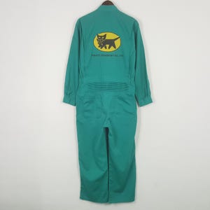 Może przedstawiać: Zielony kombinezon z długimi rękawami i kołnierzem. Z tyłu znajduje się żółty owal z czarnym logo kota i napisem "Yamato Transport Co., LTD." Kombinezon ma kieszenie i plisowany detal w talii.