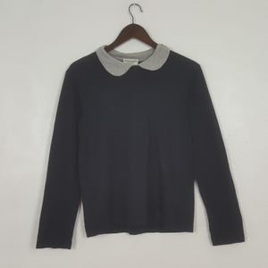 K&ouml;nnte beinhalten: Ein schwarzes Langarmshirt mit grauem Peter-Pan-Kragen. Das Kleidungsst&uuml;ck h&auml;ngt an einem Holzkleiderb&uuml;gel. Der Kragen hat ein kleines Etikett mit Text.