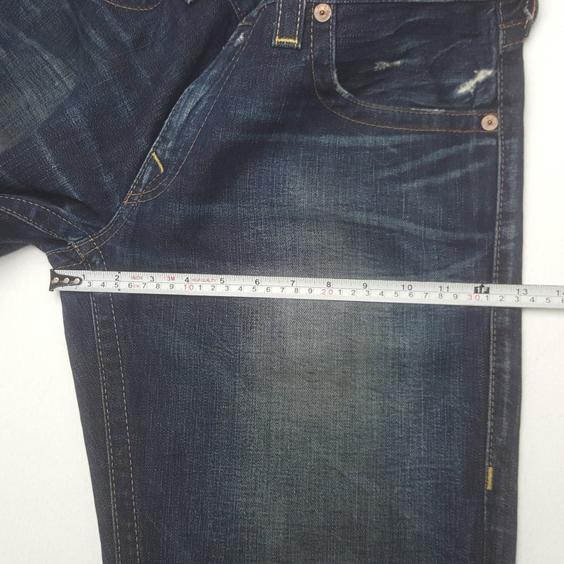 May include: Close-up of blue denim jeans with a measuring tape showing a length of approximately 33 cm. The jeans have a classic five-pocket design, with visible stitching and metal rivets. The denim has a faded, worn appearance.