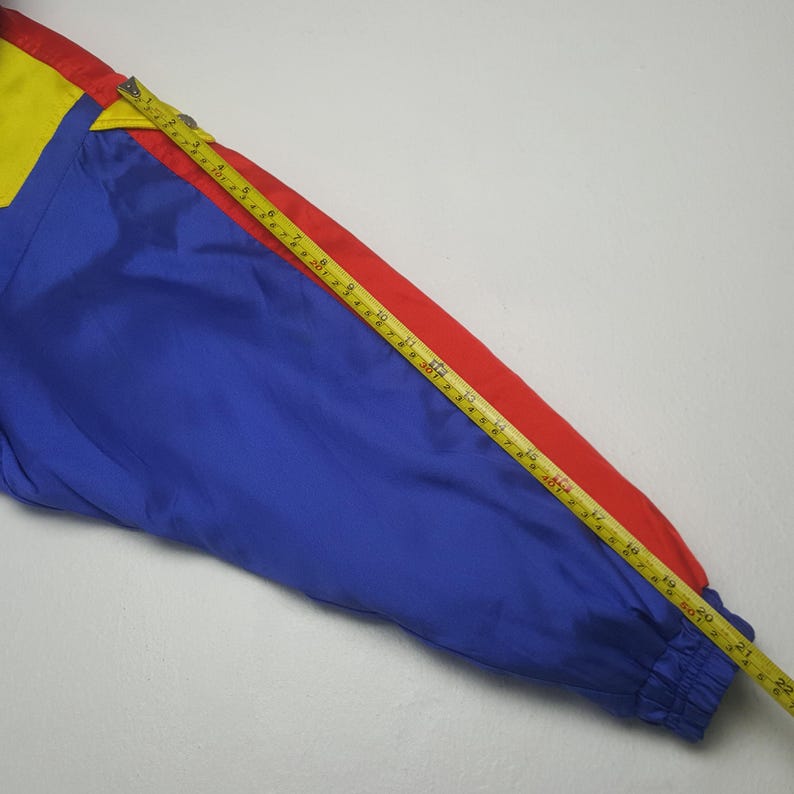 May include: A section of a colorful jacket in primary colors: blue, red, and yellow. A yellow and gold measuring tape is laid across the jacket, showing a measurement of approximately 22 inches.