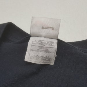 Peut inclure: Un t-shirt noir avec une &eacute;tiquette blanche portant le logo Nike. L'&eacute;tiquette indique que le t-shirt est en 100% coton et fabriqu&eacute; aux &Eacute;tats-Unis. L'&eacute;tiquette comprend &eacute;galement des informations sur la taille.