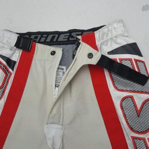 Peut inclure: Gros plan sur un pantalon de moto avec un design blanc, rouge et noir. Le pantalon a un panneau avant beige avec une fermeture &eacute;clair et des boutons-pression. La ceinture porte la marque "Dainese".