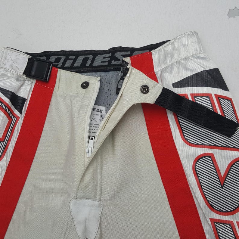 Peut inclure: Gros plan sur un pantalon de moto avec un design blanc, rouge et noir. Le pantalon a un panneau avant beige avec une fermeture &eacute;clair et des boutons-pression. La ceinture porte la marque "Dainese".