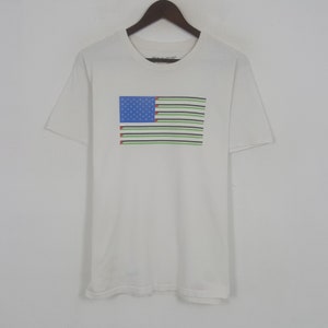 May include: White short-sleeved t-shirt featuring a graphic of a stylized American flag. The flag design includes a blue rectangle with white stars, horizontal stripes in green, black, and white, and red accents.