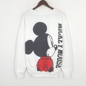 Może przedstawiać: Biała bluza z długim rękawem z dużą grafiką Myszki Miki w kolorach czarnym, czerwonym i białym. Słowa "MICKEY MOUSE" są wydrukowane pionowo na prawym rękawie. Bluza wisi na drewnianym wieszaku.